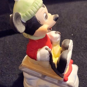 May include: A ceramic Mickey Mouse figurine, wearing a red jacket, green hat, and white gloves, on a wooden sled with red details. The sled sits on a snowy base. This collectible figurine is a winter-themed item.