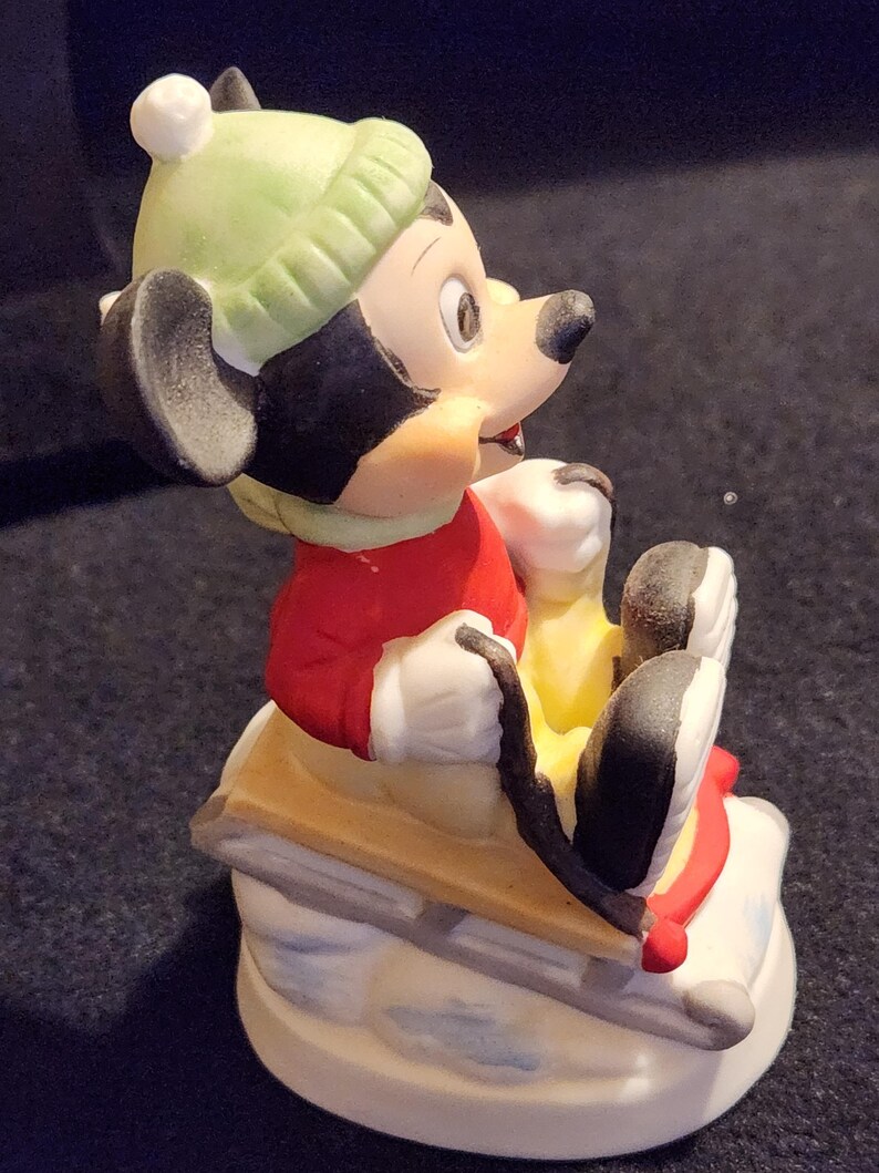 May include: A ceramic Mickey Mouse figurine, wearing a red jacket, green hat, and white gloves, on a wooden sled with red details. The sled sits on a snowy base. This collectible figurine is a winter-themed item.