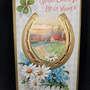 May include: Vintage postcard with a gold horseshoe, a landscape scene, and the text "Good Luck and Best Wishes." The card features a four-leaf clover, white daisies, and blue forget-me-nots. The background has a plaid pattern.