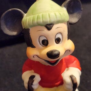 May include: A Mickey Mouse figurine wearing a light green knitted hat and a red top. The figurine has black ears, a black nose, and yellow trousers. The figure is smiling and has white gloves.