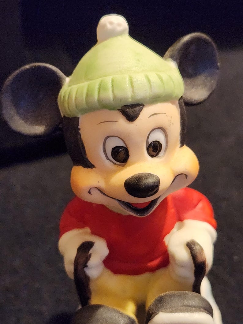 May include: A Mickey Mouse figurine wearing a light green knitted hat and a red top. The figurine has black ears, a black nose, and yellow trousers. The figure is smiling and has white gloves.