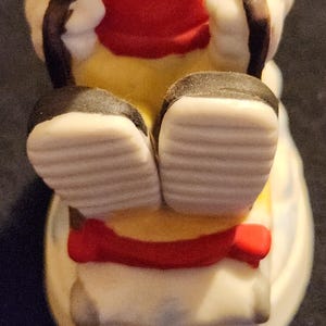 May include: A ceramic figurine of a seated character, viewed from the feet up. The figure wears a red coat, yellow trousers, and black shoes with white soles. A red bow is visible. The figurine is on a white base.