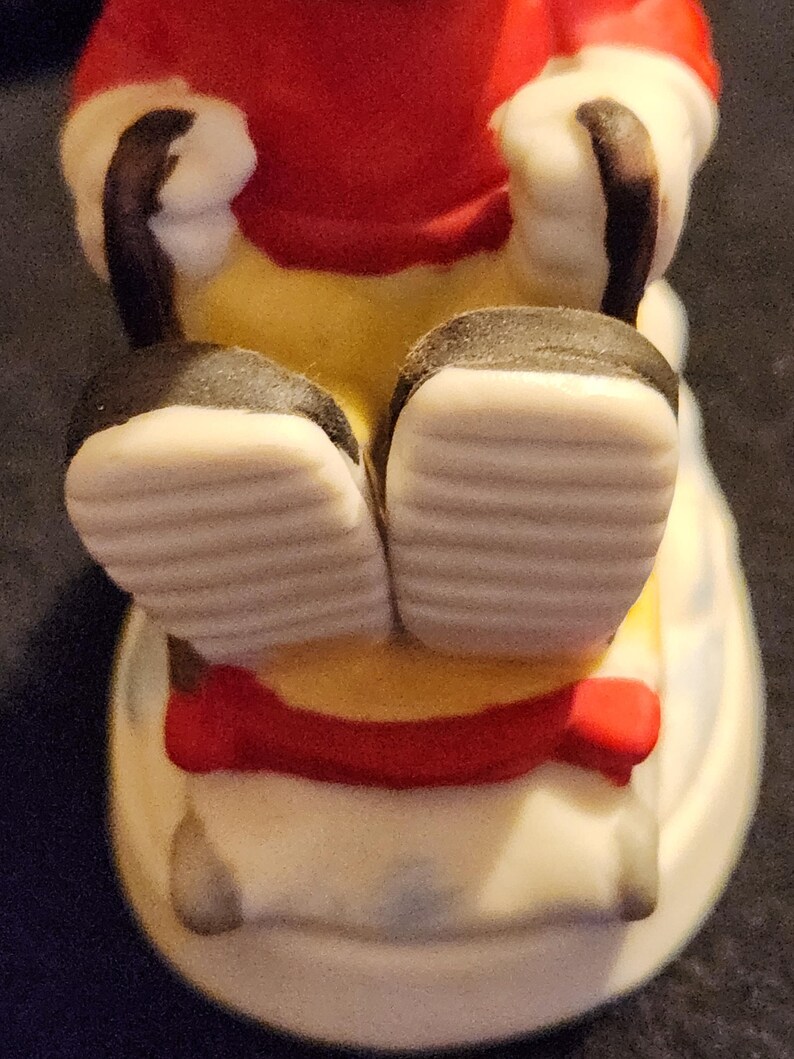 May include: A ceramic figurine of a seated character, viewed from the feet up. The figure wears a red coat, yellow trousers, and black shoes with white soles. A red bow is visible. The figurine is on a white base.