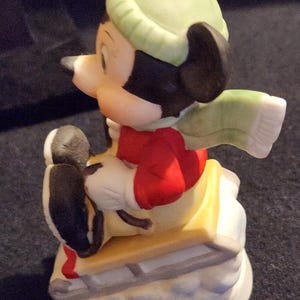 May include: A ceramic Mickey Mouse figurine on a sled. Mickey is dressed in a red top, green hat, and scarf. The sled is yellow and white, and the base is white with blue details. A collectible item.