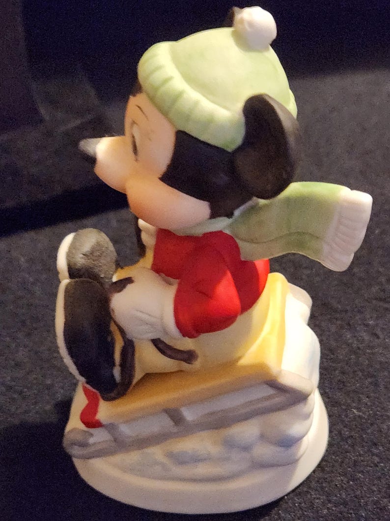 May include: A ceramic Mickey Mouse figurine on a sled. Mickey is dressed in a red top, green hat, and scarf. The sled is yellow and white, and the base is white with blue details. A collectible item.