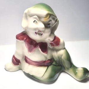 May include: A ceramic figurine of a seated elf with a green hat and pants, and a white shirt with red accents. The elf is smiling with its hand resting on its cheek. The figurine has a vintage, hand-painted appearance.