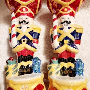 May include: Two decorative nutcracker figurines. Each figurine is painted with a red hat, gold trim, and a white face with a black mustache. They wear blue and gold uniforms and hold a red and white striped candy cane. The base is yellow with red accents.