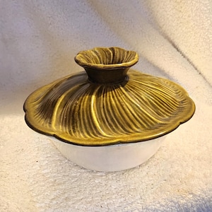 May include: A ceramic serving bowl with a lid. The lid is a golden-brown color with a textured, ridged design. The bowl is white with a dark brown rim. The lid has a decorative handle.