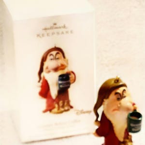May include: A Grumpy dwarf figurine from Snow White holding a dark green mug, positioned in front of a white Hallmark Keepsake box. The figurine wears a brown hat and a red tunic, with a long white beard and a stern facial expression.