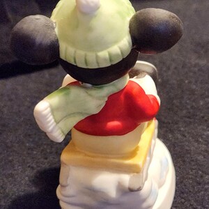 May include: A ceramic figurine of a cartoon character in winter clothing. The figure is wearing a red jacket, a light green hat, and a matching scarf. The figurine is on a snowman base with a yellow platform.