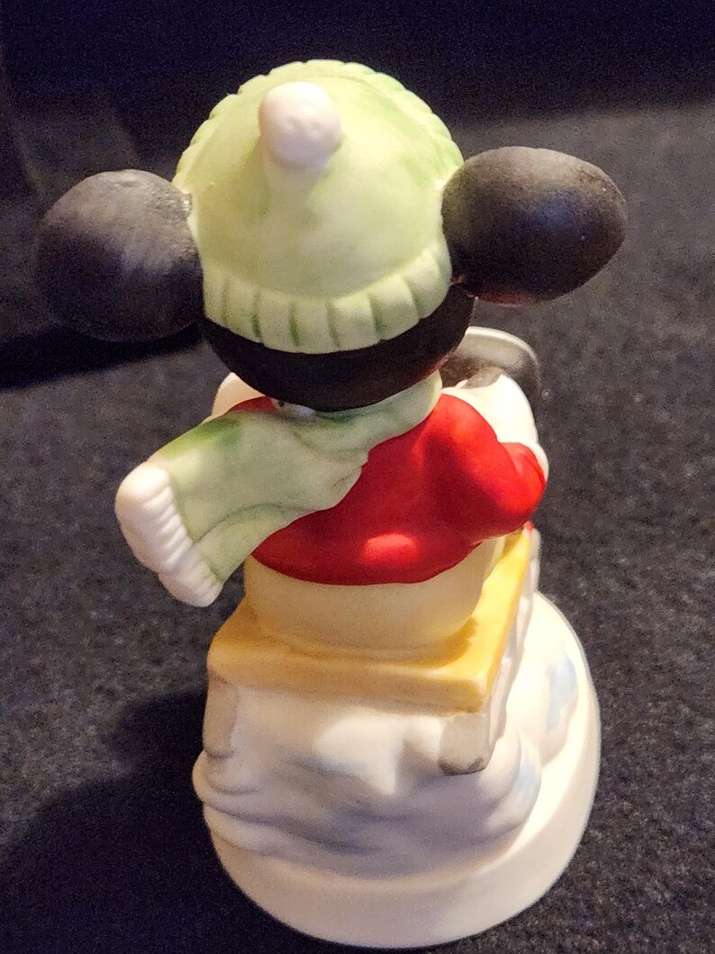 May include: A ceramic figurine of a cartoon character in winter clothing. The figure is wearing a red jacket, a light green hat, and a matching scarf. The figurine is on a snowman base with a yellow platform.