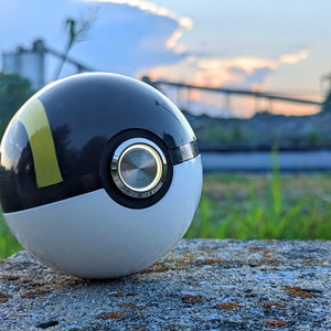 May include: A black and white Poke Ball with a yellow stripe and a silver button. The Poke Ball is sitting on a gray rock.