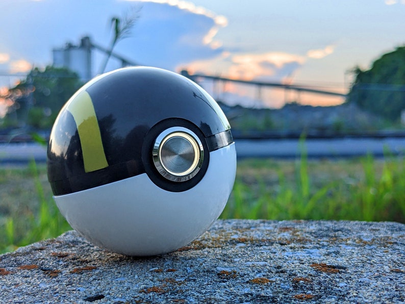 May include: A black and white Poke Ball with a yellow stripe and a silver button. The Poke Ball is sitting on a gray rock.