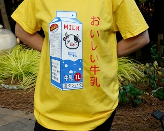 Könnte beinhalten: Gelbes T-Shirt mit einem Cartoon-Milchkarton-Design und einer Kuh-Illustration. Auf dem Karton steht "MILK" und japanischer Text. Das Shirt hat einen Rundhalsausschnitt.