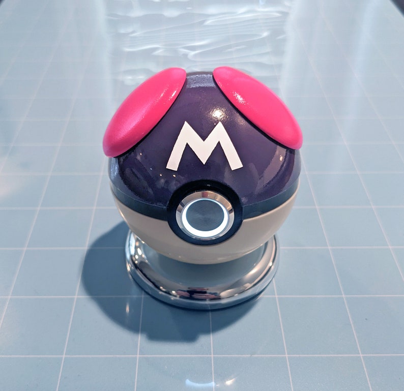 May include: A purple and white Poke Ball with a pink button on top and the letter "M" on the front.