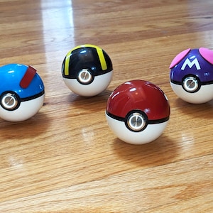 May include: Four Poke Balls with different colored designs. The Poke Balls are red, blue, black, and purple. Each Poke Ball has a white button in the center.