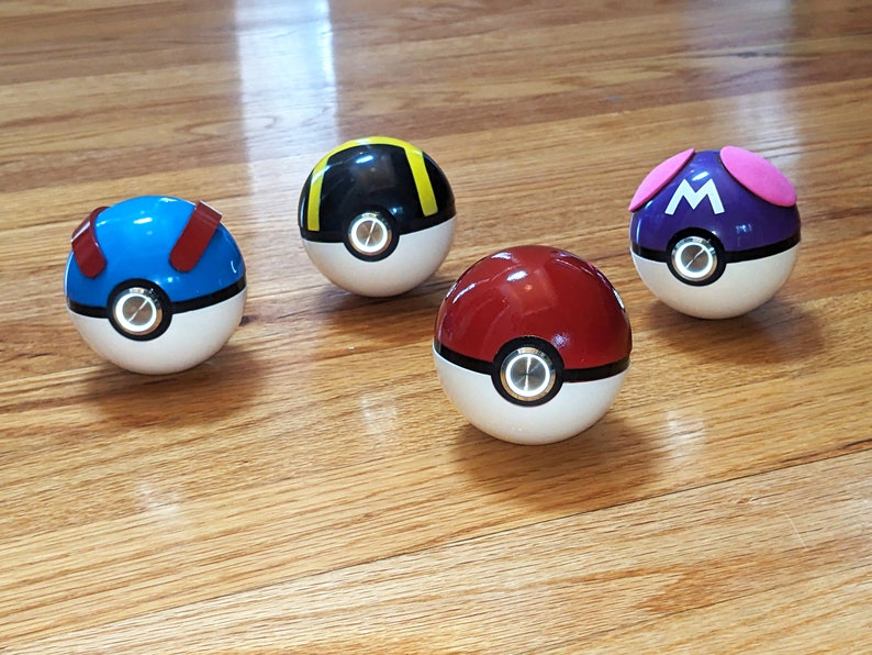 May include: Four Poke Balls with different colored designs. The Poke Balls are red, blue, black, and purple. Each Poke Ball has a white button in the center.