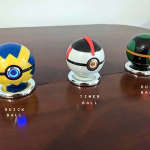 May include: Three Poke Balls, a yellow and blue Quick Ball, a white Timer Ball, and a green and black Dusk Ball. Each ball has a button on the front and is sitting on a silver base.