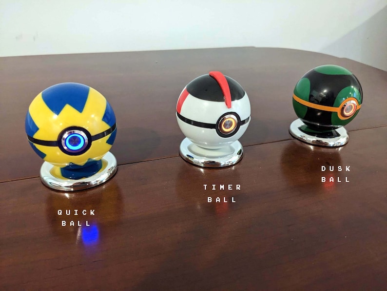 May include: Three Poke Balls, a yellow and blue Quick Ball, a white Timer Ball, and a green and black Dusk Ball. Each ball has a button on the front and is sitting on a silver base.
