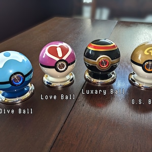 May include: Four different Poke Balls, each with a unique design and color. The Poke Balls are on a wooden surface. The Poke Balls are labeled "Dive Ball", "Love Ball", "Luxury Ball", and "G.S. Ball".