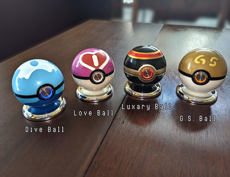 May include: Four different Poke Balls, each with a unique design and color. The Poke Balls are on a wooden surface. The Poke Balls are labeled "Dive Ball", "Love Ball", "Luxury Ball", and "G.S. Ball".