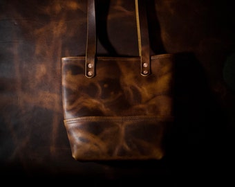 May include: A brown leather tote bag with two straps and copper rivets. The bag has a distressed look and is made of high-quality leather.