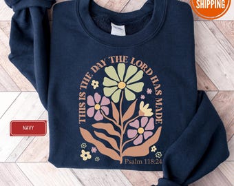 May include: Navy blue sweatshirt with a floral design in pastel colors. The design includes the text "THIS IS THE DAY THE LORD HAS MADE" and "Psalm 118:24". The sweatshirt has a crew neck and long sleeves.