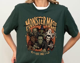 May include: A dark green short-sleeve t-shirt featuring a graphic print of classic monsters emerging from a large orange pumpkin. The text reads MONSTER MASH and EST. 1962 in a stylized font. The design includes a werewolf, Frankenstein, a witch, a ghost, and a vampire.