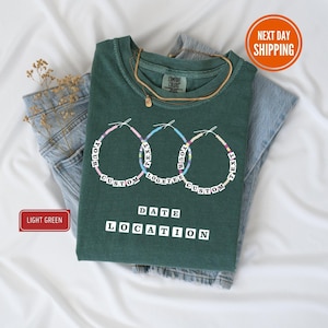 Könnte beinhalten: Ein hellgrünes T-Shirt mit einem individuellen Design mit drei Perlenkreisen mit den Worten "YOUR CUSTOM TEXT" darum. Unter den Kreisen stehen die Worte "DATE LOCATION". Das Shirt liegt gefaltet auf einer Jeans.
