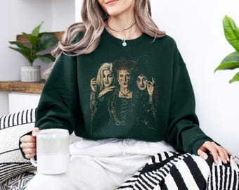 May include: A dark forest green crewneck sweatshirt featuring a vintage-style graphic print of three women with distinct hairstyles and period-style clothing. The central figure has red hair, while the others have blonde and dark hair.