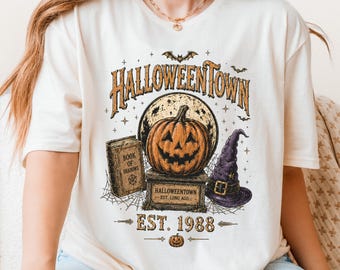 May include: A cream-colored t-shirt featuring a graphic print with the text HALLOWEENTOWN EST. 1988. The design includes a jack-o'-lantern on a pedestal, a book titled BOOK OF SHADOWS, a purple witch hat, bats, and spiderwebs.