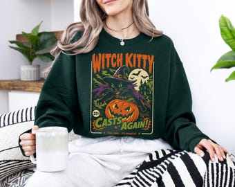May include: A dark green crewneck sweatshirt featuring a graphic print of a black cat wearing a witch hat, a jack-o'-lantern, and the text Witch Kitty Casts Again! This time... it's magical. The design includes a full moon and bats.