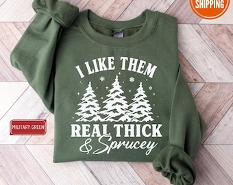 May include: A military green sweatshirt with white text and graphics. The text reads "I LIKE THEM REAL THICK & Sprucey" above three stylized Christmas trees. The sweatshirt is folded on a light wood surface.
