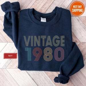 May include: Navy blue sweatshirt with the text "VINTAGE 1980" in a retro font. The text is in a variety of colors, including yellow, teal, red, and pink. The sweatshirt has a crew neck and long sleeves. The word "NAVY" is in a red box.