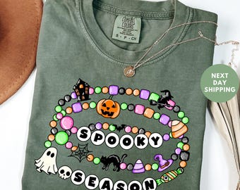 May include: Olive green t-shirt with a colorful Halloween-themed graphic. The design includes a pumpkin, ghost, witch, and the words "Spooky Season". A silver necklace with a pendant is draped across the shirt.