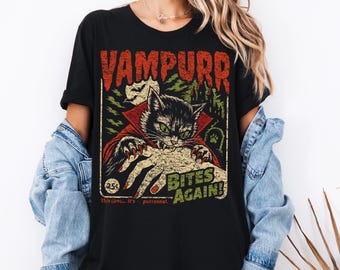 May include: A black t-shirt featuring a graphic print of a vampire cat with red eyes and fangs, wearing a red cape. The text reads VAMPURR BITES AGAIN! with a 25 cent comic book style badge, a moon, and a castle silhouette in the background.
