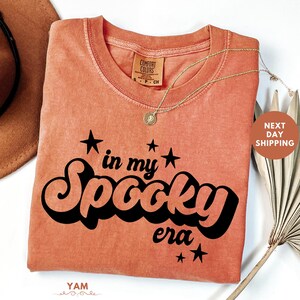 May include: Orange Comfort Colors t-shirt with the phrase "in my Spooky era" in black script and stars. A gold necklace with a pendant is draped on the shirt. A brown hat and dried palm leaf are in the background.
