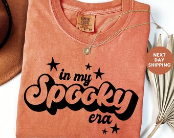 May include: Orange Comfort Colors t-shirt with the phrase "in my Spooky era" in black script and stars. A gold necklace with a pendant is draped on the shirt. A brown hat and dried palm leaf are in the background.