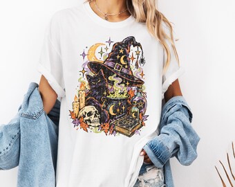 May include: A white t-shirt featuring a graphic print of a black cat, a witch hat, a bubbling cauldron, a skull, candles, a spellbook, and a crescent moon. The design includes stars, autumn leaves, and a spider hanging from the hat.