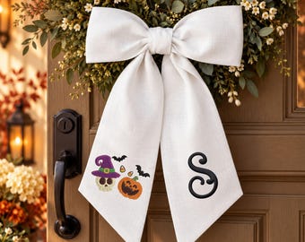 May include: A large white fabric bow featuring two long tails. One tail displays an embroidered skull wearing a purple witch hat, a jack-o'-lantern, candy corn, and black bats. The other tail features a large, black embroidered letter S.