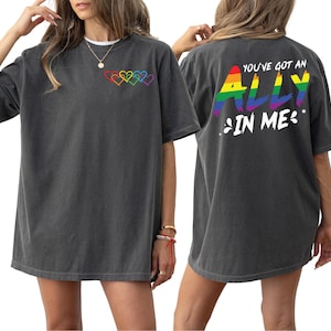 May include: Dark gray t-shirt with rainbow heart design on the front and the text "YOU'VE GOT AN ALLY IN ME" on the back. The shirt is a short-sleeved, unisex style.