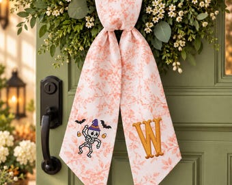 May include: A fabric door hanger featuring a light pink and white floral toile pattern. The left tail displays an embroidered skeleton wearing a purple witch hat with bats and candy corn, while the right tail features a large, gold embroidered letter W.
