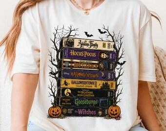 May include: A cream-colored t-shirt featuring a graphic of a stack of seven VHS tapes titled The Addams Family, Hocus Pocus, Beetlejuice, Halloweentown, Halloweentown II, The Nightmare Before Christmas, Goosebumps, and The Witches.