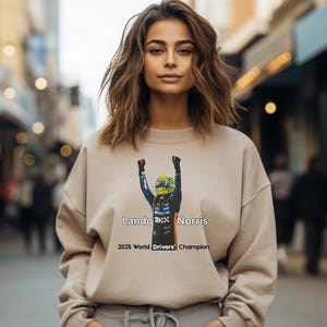May include: Beige sweatshirt featuring a graphic of a Formula 1 driver with the text "Lando Norris" and "2025 World Drivers' Champion". The design shows the driver in a racing suit with arms raised. The sweatshirt has a drawstring and is worn by a person.
