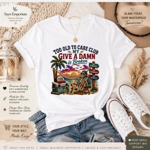 May include: A white t-shirt featuring a graphic of a skeleton relaxing in a beach chair under a palm tree with a hammock, a cooler, and signs reading No Schedule, No Reminders, and No Regrets, with text stating Too Old To Care Club My Give A Damn Is Broken.