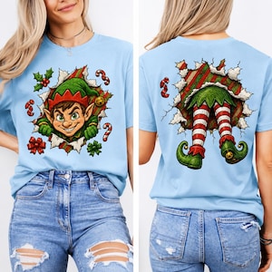 May include: A light blue t-shirt featuring a graphic of a Christmas elf. The front shows the elf's face peeking through a torn paper effect, while the back displays the elf's striped legs and curled shoes emerging from a similar torn opening.