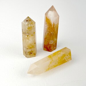 Pode incluir: Três pontas de cristal de citrino amarelo sobre um fundo branco. Os cristais são transparentes com um leve tom amarelo e têm uma ponta pontiaguda.
