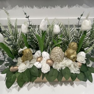 Puede incluir: Un centro de mesa decorativo de Pascua con tulipanes blancos, vegetación artificial y detalles dorados. El arreglo incluye figuras de conejos dorados, huevos moteados y un huevo dorado texturizado, todo en una caja de madera.