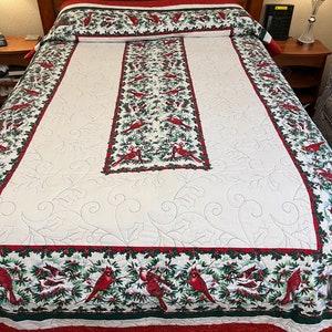 Puede incluir: Un edredón blanco acolchado con un borde rojo, verde y blanco. El borde presenta un patrón repetido de cardenales, acebo y bayas. El centro del edredón es blanco con un patrón acolchado.