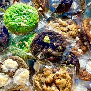 May include: An assortment of freshly baked cookies in clear plastic packaging. The cookies vary in color, including green, brown, and purple, and feature toppings like white chocolate chips, marshmallows, and nuts. A variety of textures and flavors are visible.
